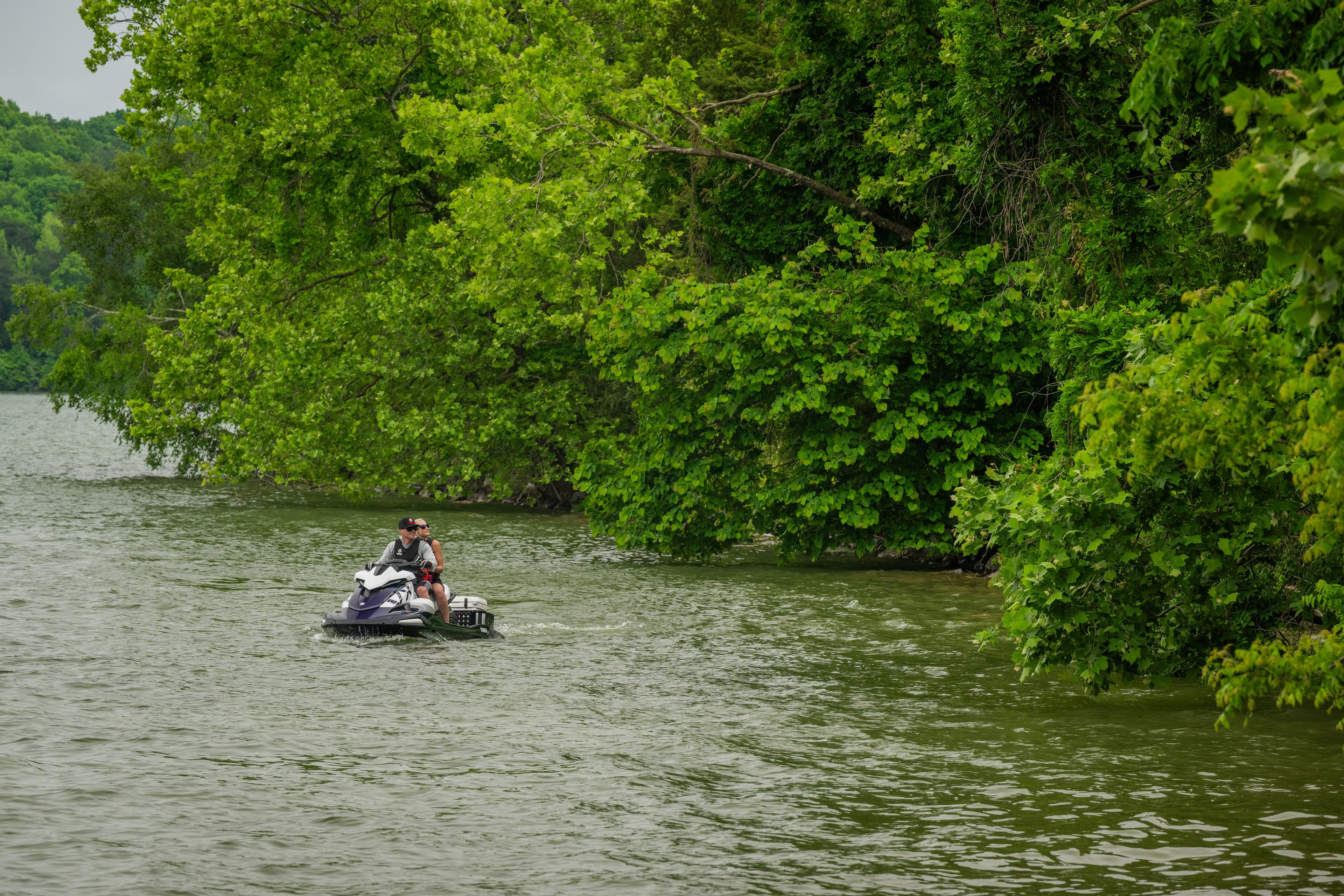 White Yamaha jet ski with two riders on lake water near wooded shore