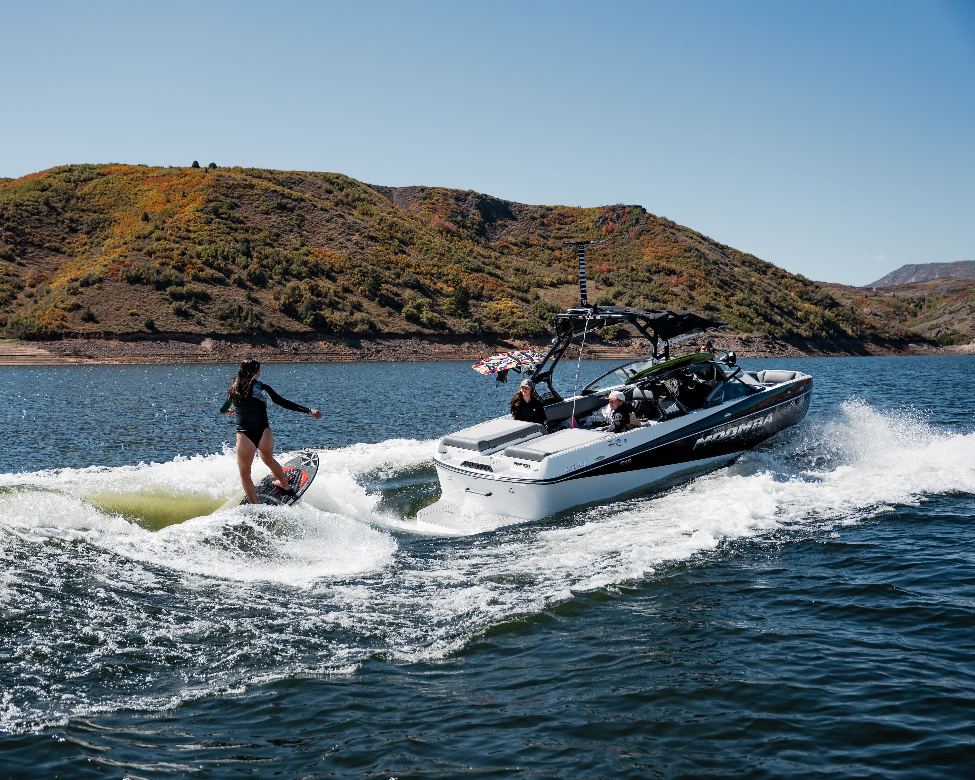 A Moomba wakeboard boat underway on water with a wakeboarder being towed behind it, piloted by an operator in the helm seat.