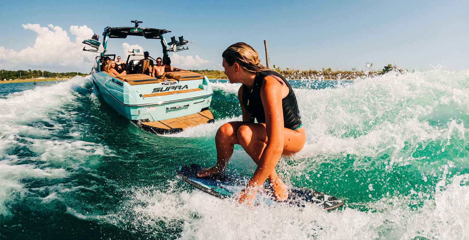 Woman wakesurfing behind a turquoise Supra wakeboard boat with wake tower, creating large wake on clear blue water
