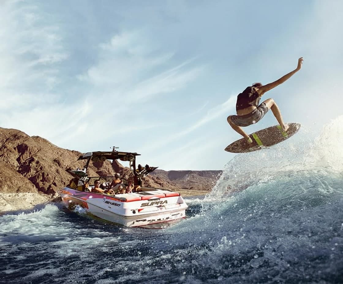 A wakeboarder launches into the air behind a white Supra boat equipped with a wakeboard tower.
