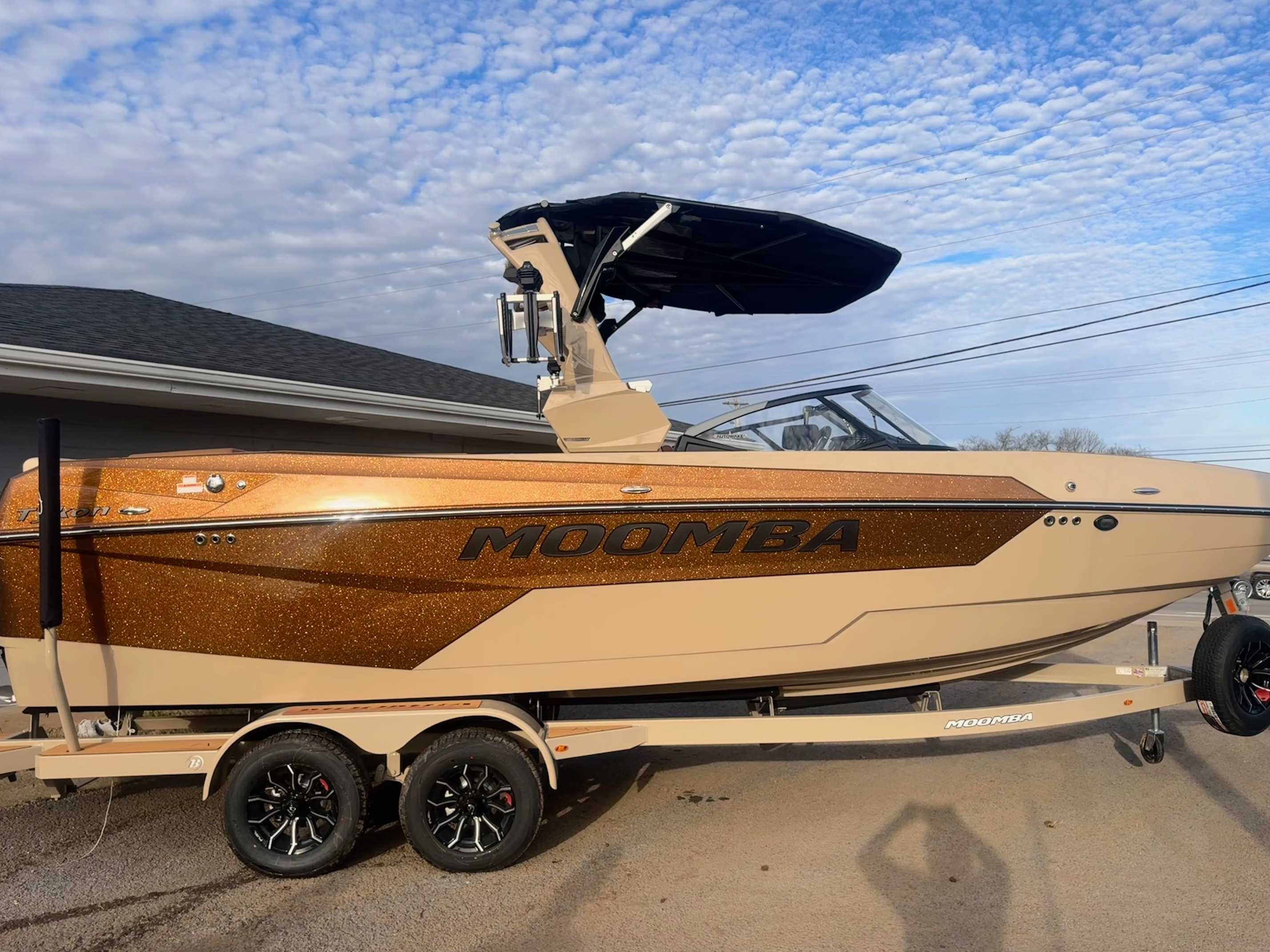 Bronze metallic Moomba wakeboard boat with folding wake tower mounted on dual-axle trailer parked in residential driveway