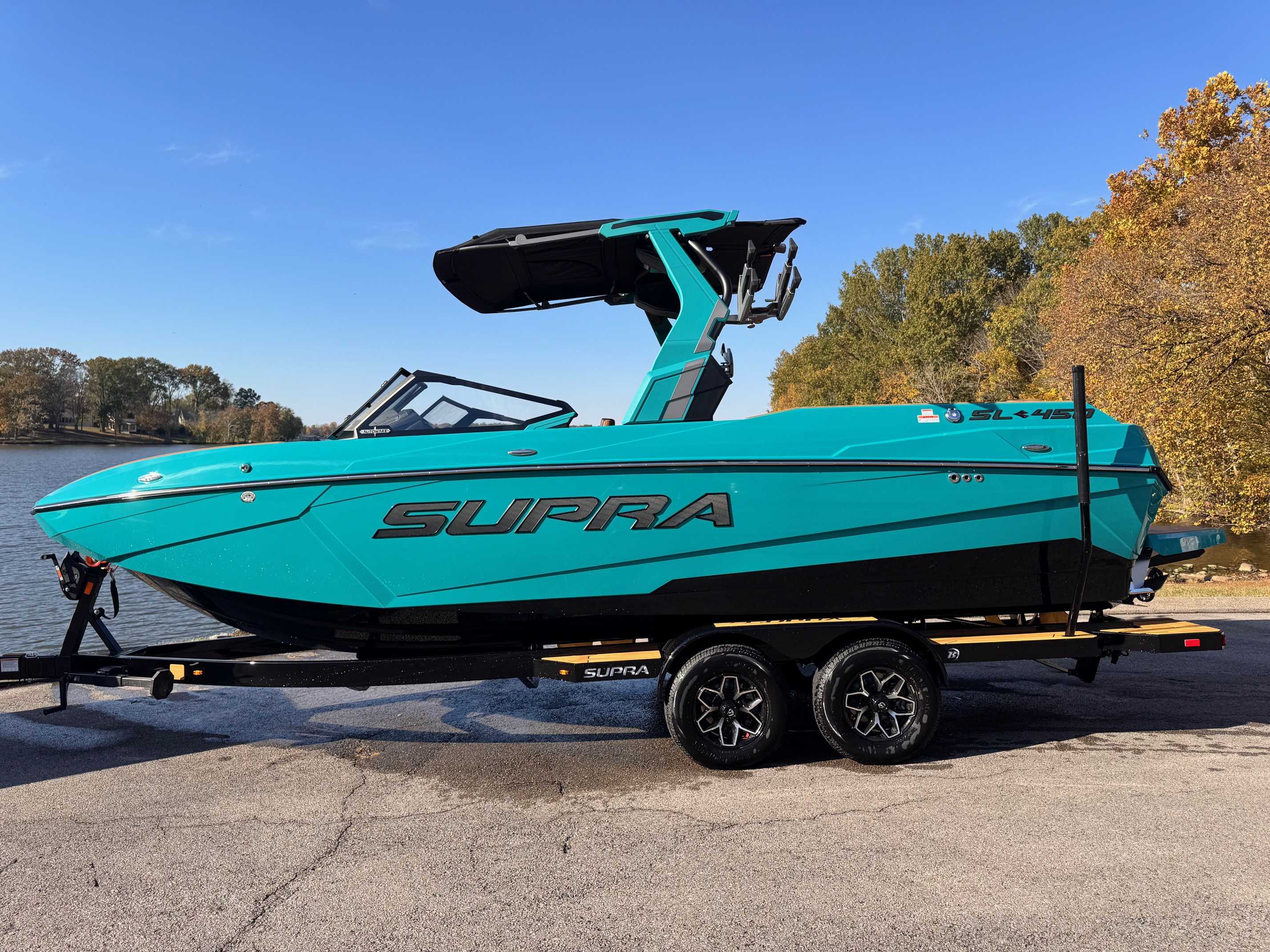 Turquoise Supra wakeboard boat with black tower on trailer at lakeside with fall foliage