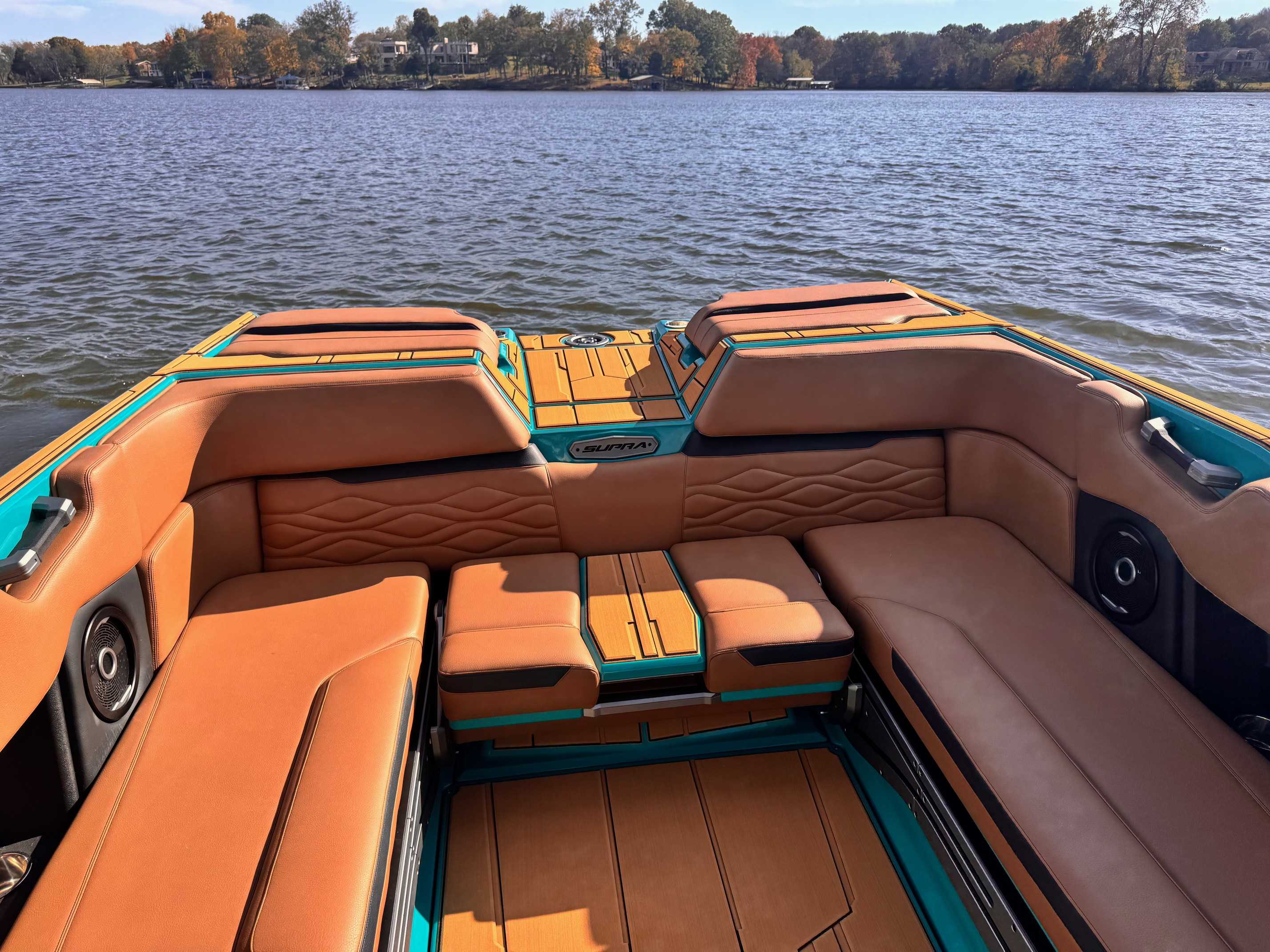 Interior cockpit view of Supra wakeboard boat featuring brown leather wraparound seating with teal trim, positioned on calm lake with fall foliage in background